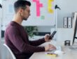 Handsome entrepreneur sending messages with smart phone while working with computer