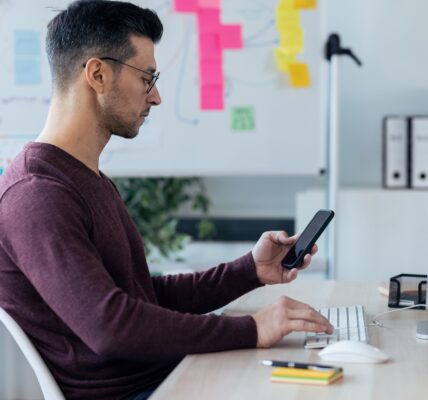 Handsome entrepreneur sending messages with smart phone while working with computer