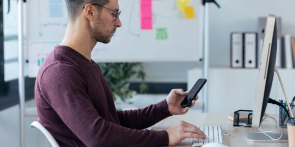 Handsome entrepreneur sending messages with smart phone while working with computer
