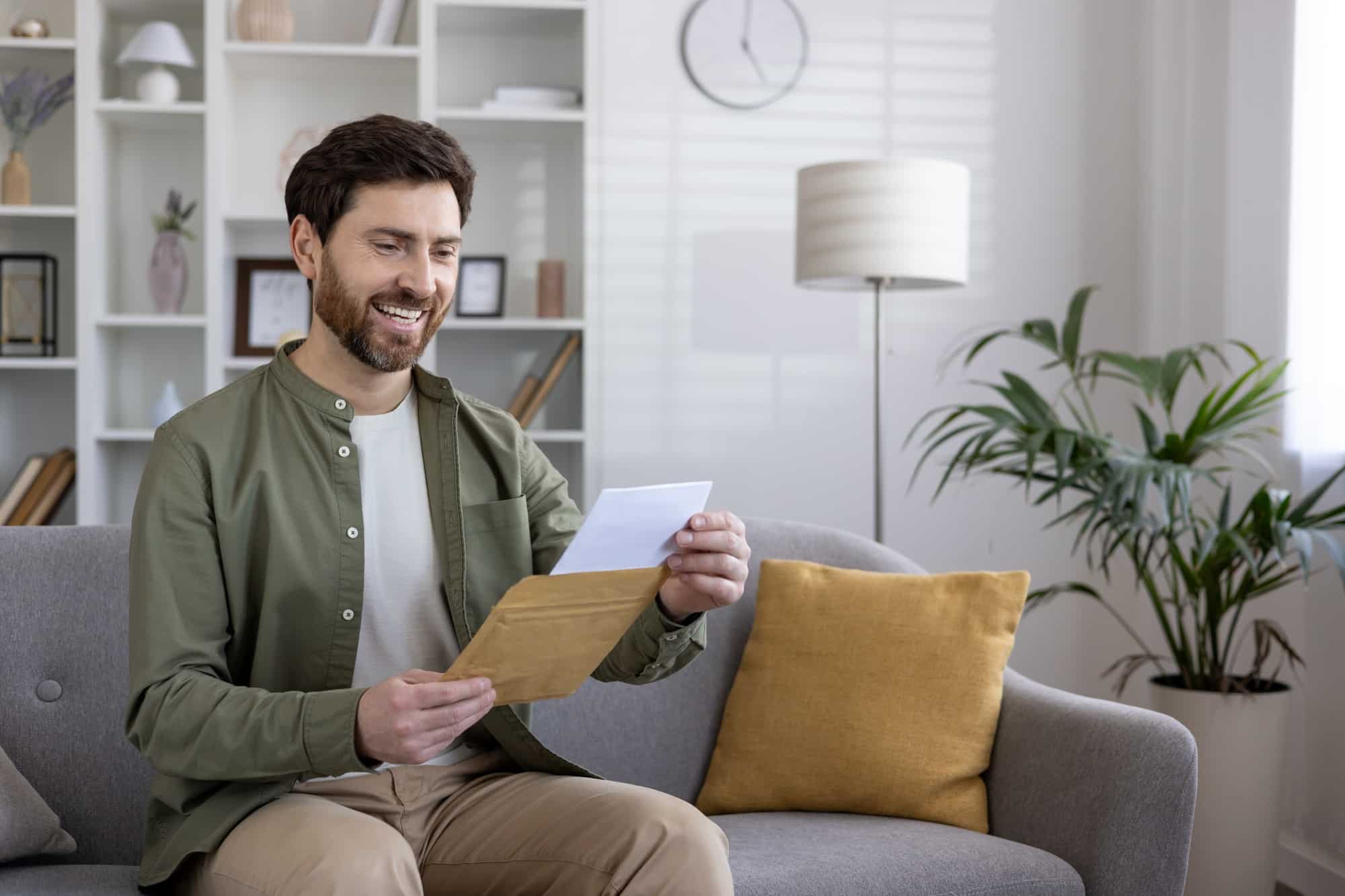 Smiling man reading letter on couch in modern living room